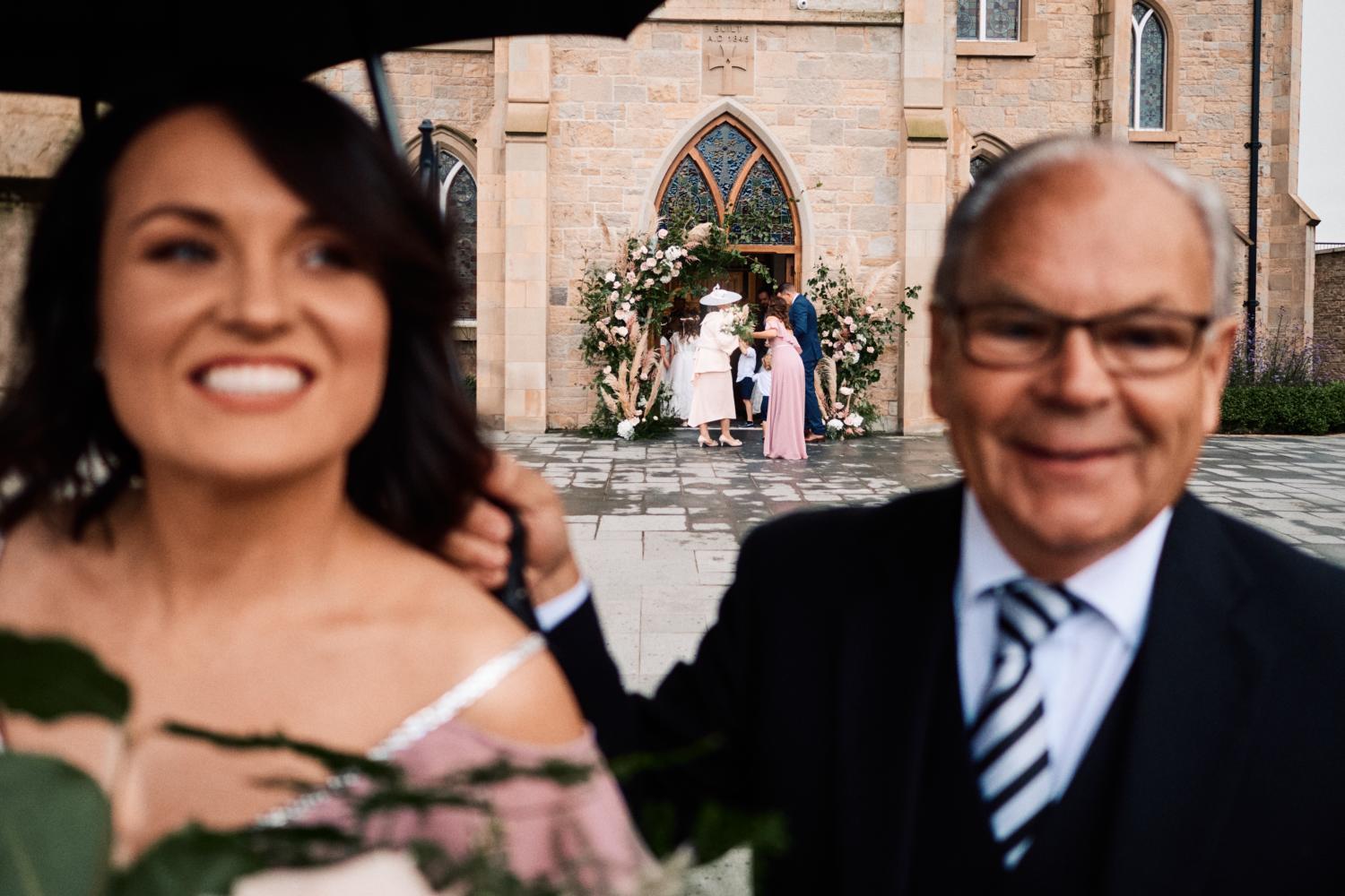 A driver holds an umbrella for a bridesmaid outside a church