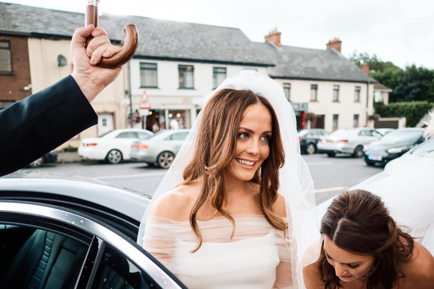 A driver holds an umbrella for a smiling bride in Dungannon