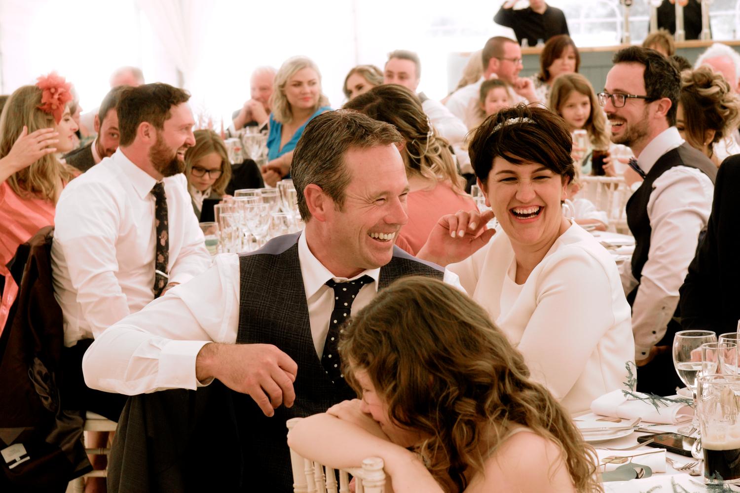 A family laugh at a table during wedding speeches in a marquee