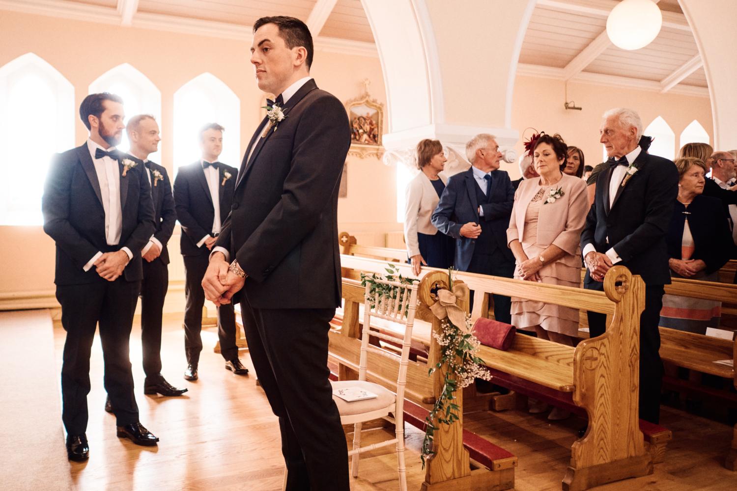 A groom in a tuxedo waits at the top of the aisle for the bride to arrive