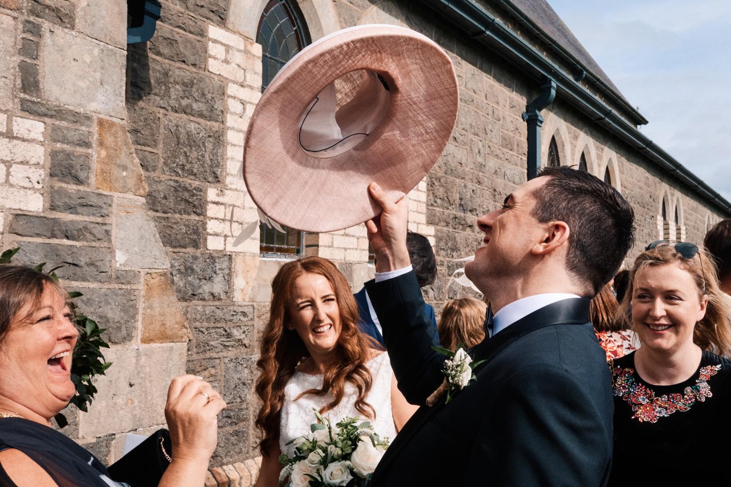 Groom lifts pink hat in the air to amusement of wedding guests