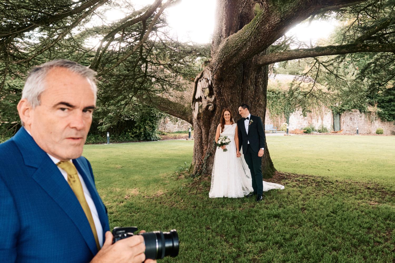 A wedding guests takes photographs of the bride and groom in front of a tree