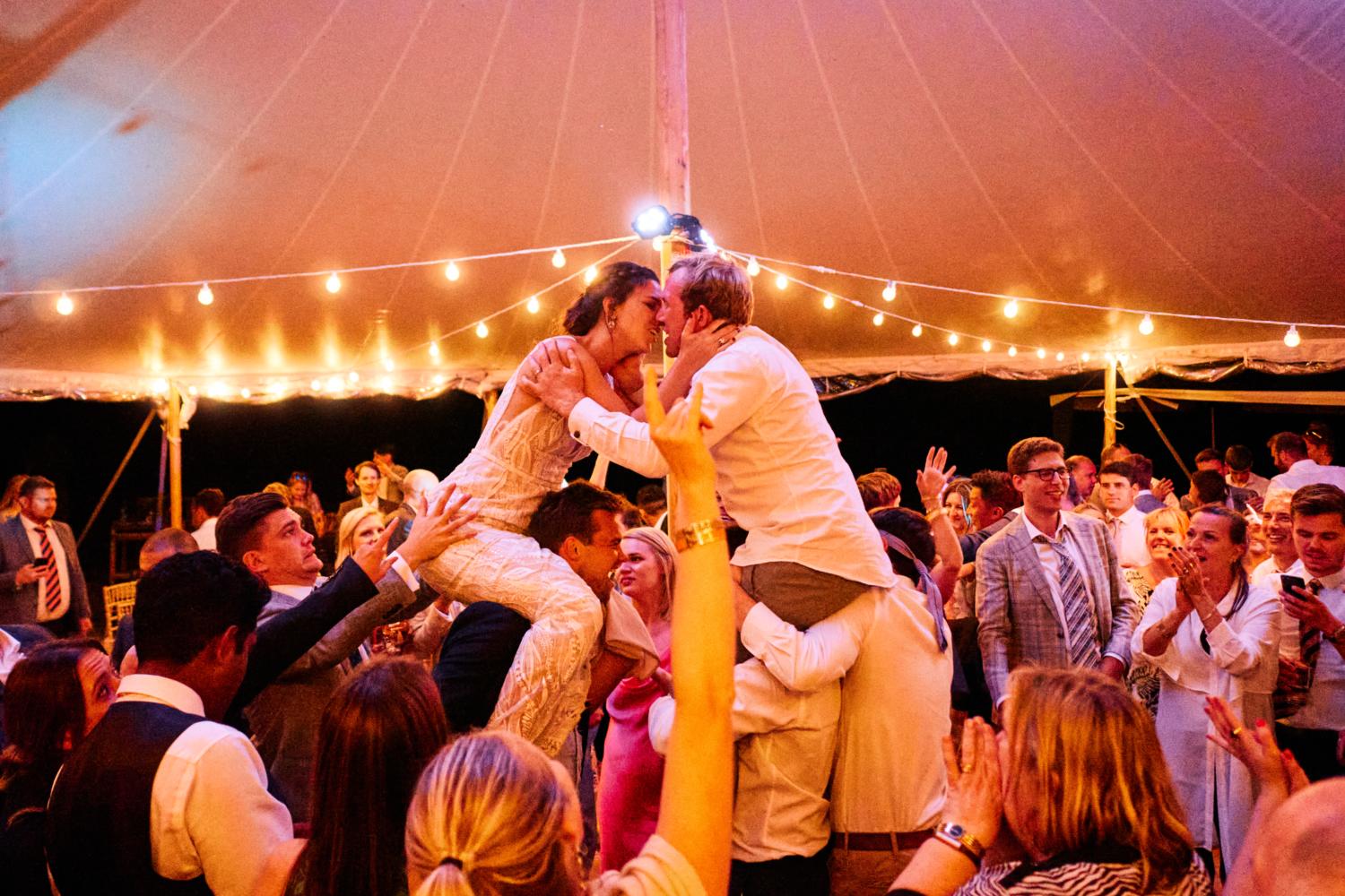 Bride and groom kiss each other on the shoulders of their friends in a marquee